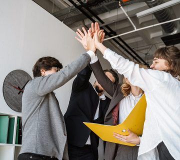 Cheerful young group of people standing in the office and giving high five 1 scaled 1 360x320